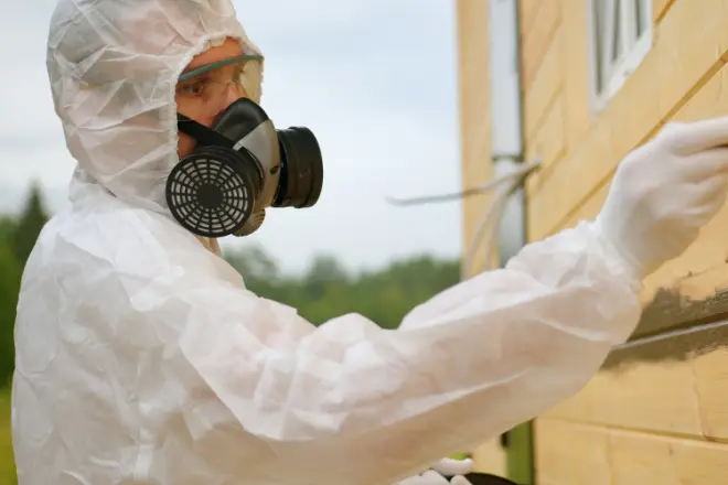 How to Remove Paint From a Home’s Exterior 1 A person in protective clothing and a respirator mask is working on the exterior of a yellow building.